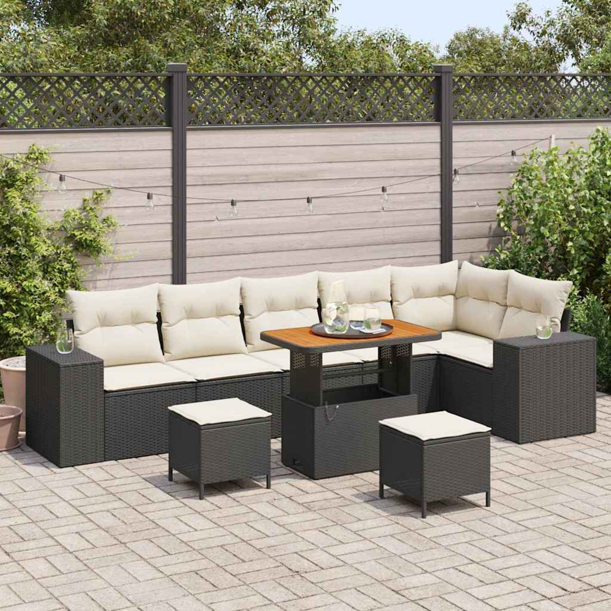 Set da patio esterno con mobili in vimini grigi e cuscini color crema. Un tavolo in legno e sgabelli sono posizionati sul patio in mattoni, con una recinzione sullo sfondo.