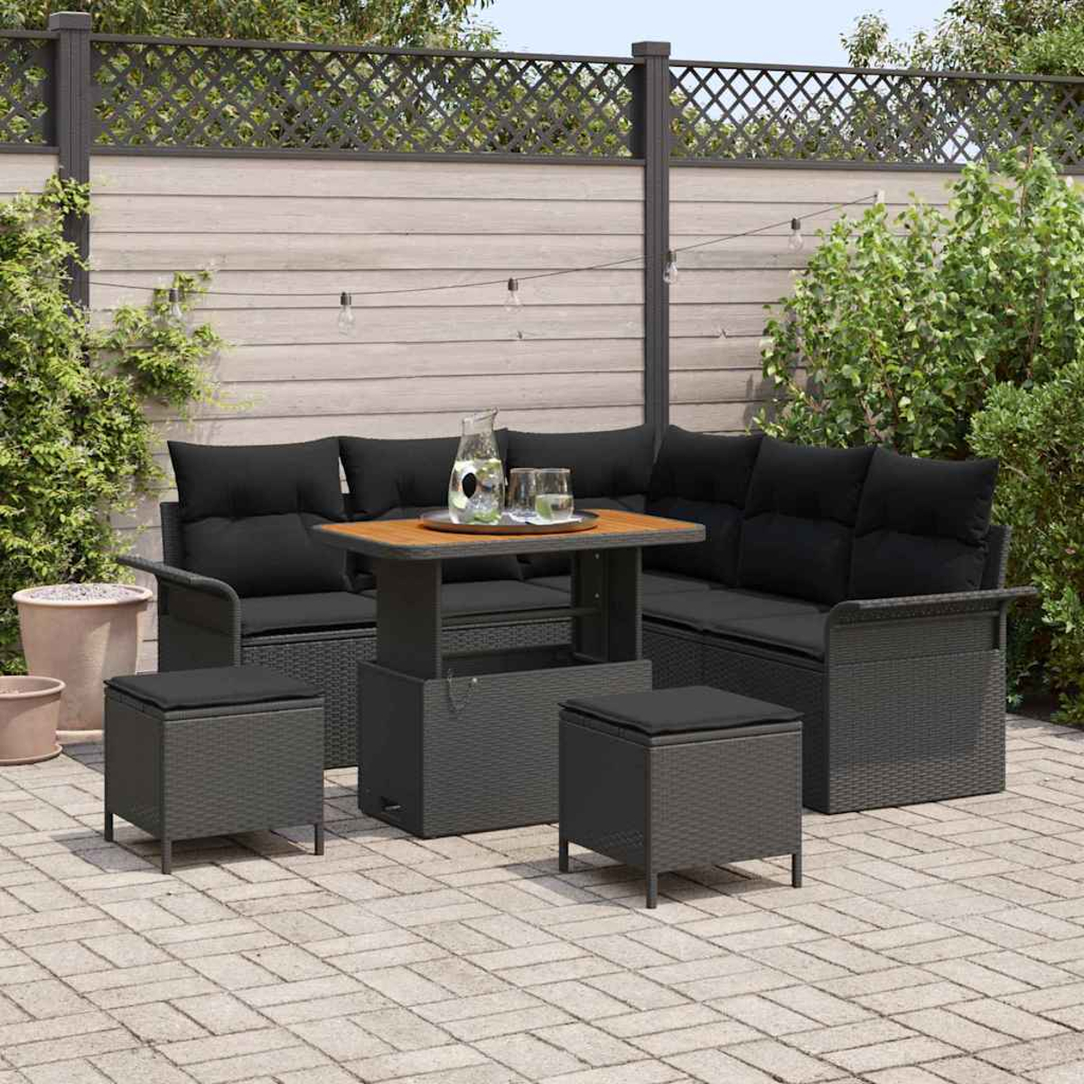 Garten-Terrassenset mit schwarzen Kissen, Tisch und Hockern auf einer Ziegelterrasse. Im Hintergrund ein Zaun und Pflanzen.