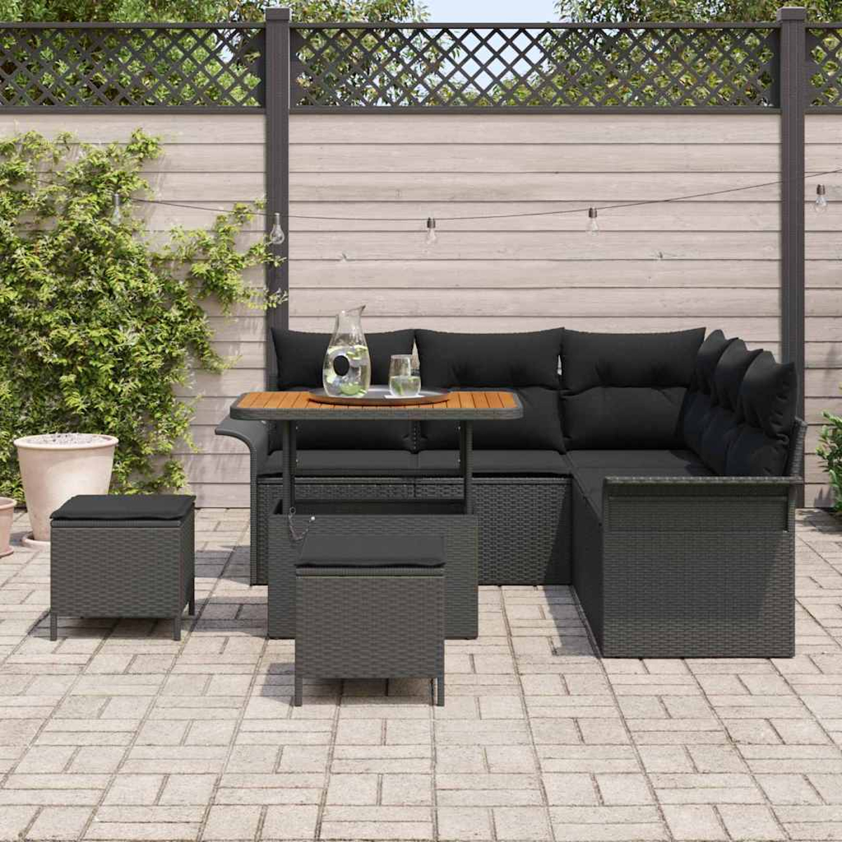 Outdoor-Ecksofa mit schwarzen Kissen und Holztisch. Zwei Würfelhocker, Wasserkrug und Gläser auf dem Tisch. Holzzaun im Hintergrund.