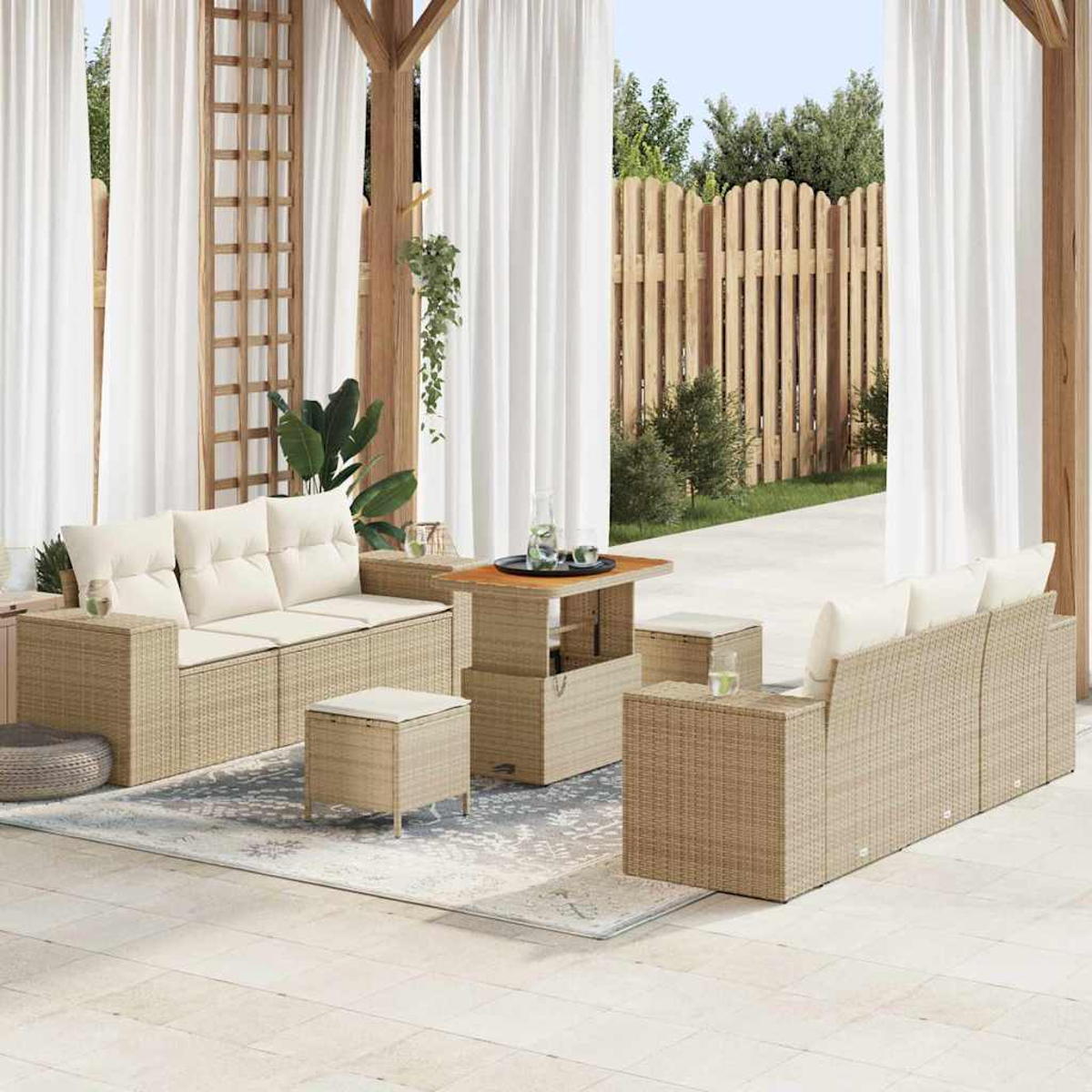 Terrassenset im Freien mit beigefarbenen Korbsitzgruppen, Ottomanen und einem Tisch. Weiße Vorhänge, Holzzaun und grüne Pflanzen.