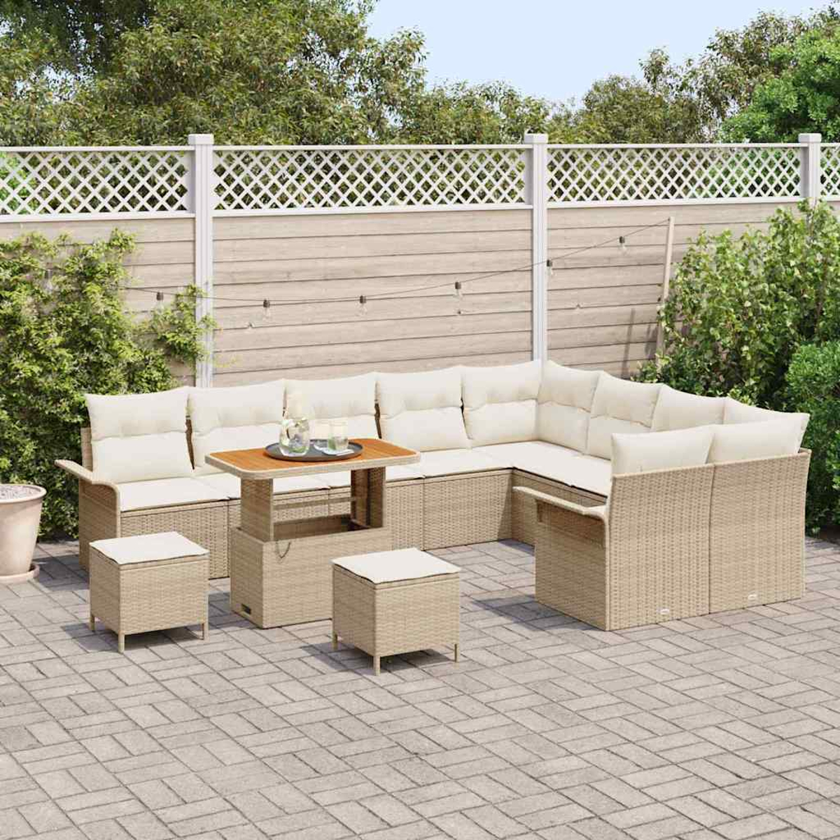 Juego de patio al aire libre: sofá seccional de mimbre beige con cojines crema, mesa de madera, dos otomanas en un patio de ladrillos, valla blanca al fondo.