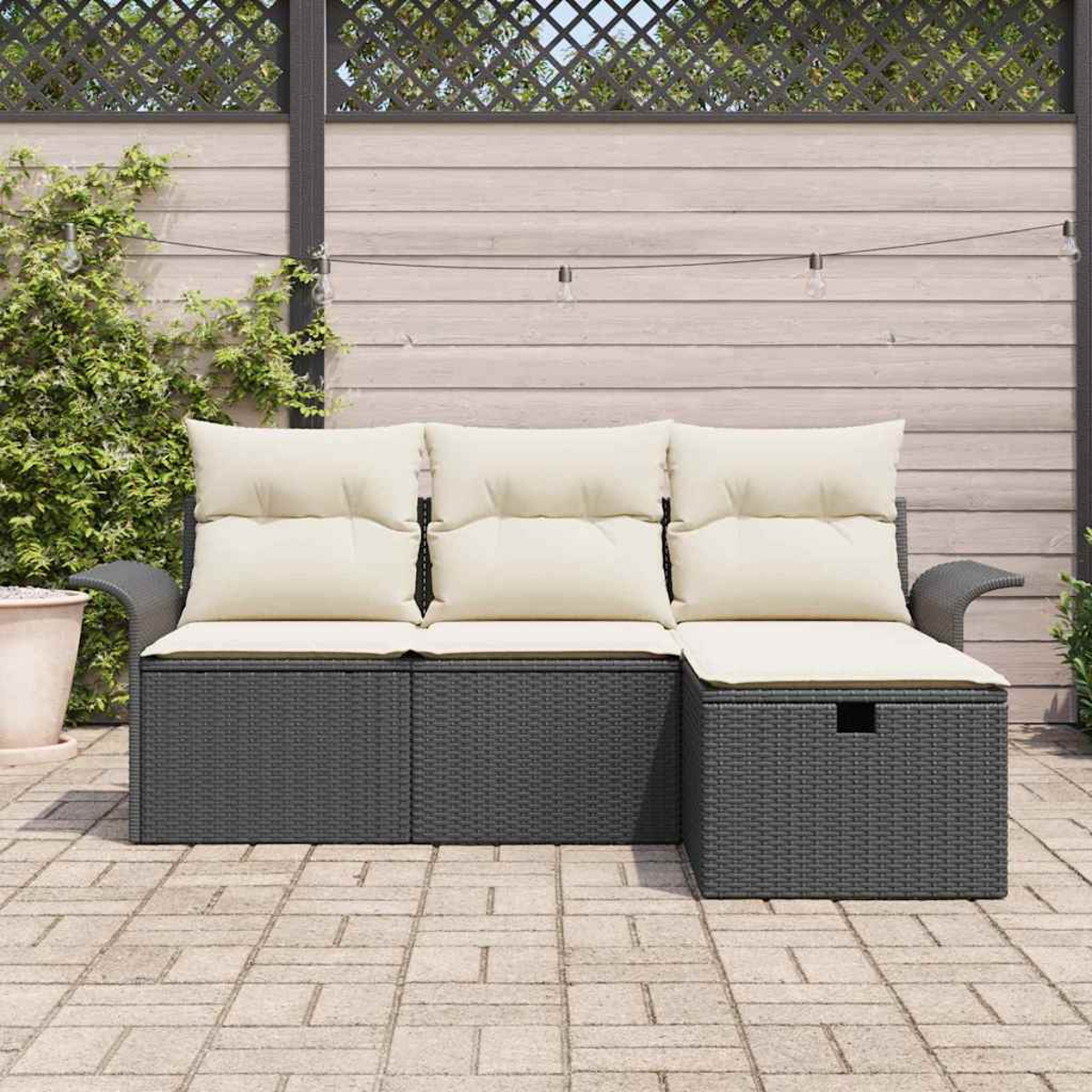 Graues Korbmöbel mit cremefarbenen Kissen auf einer Ziegelterrasse. Holzzaun und Kletterpflanzen im Hintergrund.