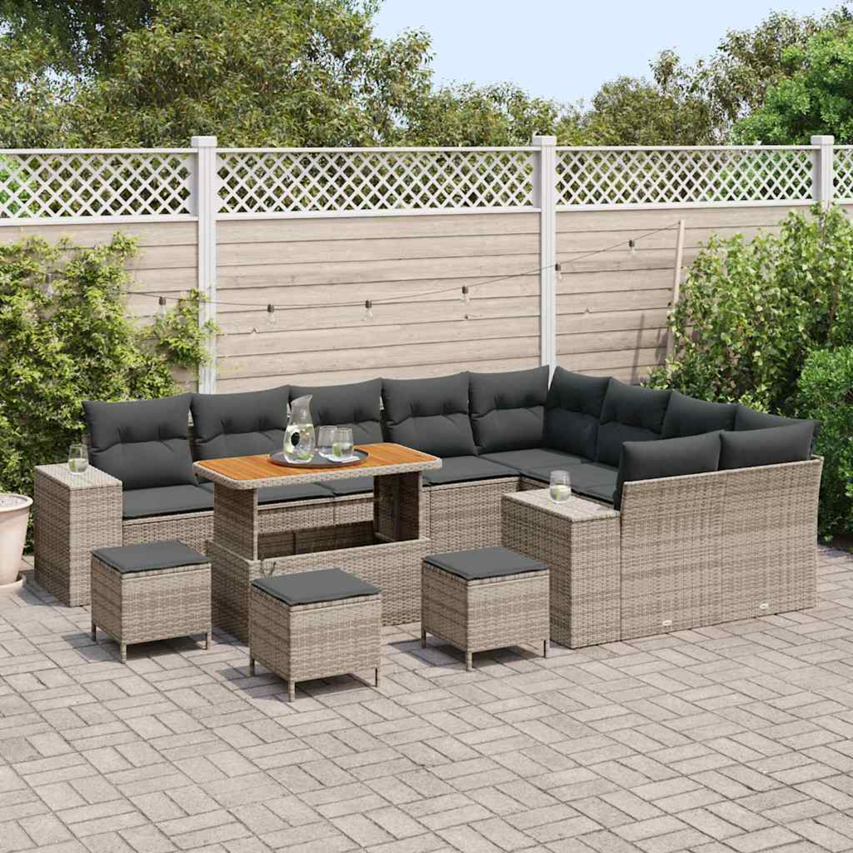 Eine Sitzgruppe im Freien mit grauen Kissen, einem Holztisch und passenden Hockern. Auf einer Ziegelterrasse mit Zaun im Hintergrund.