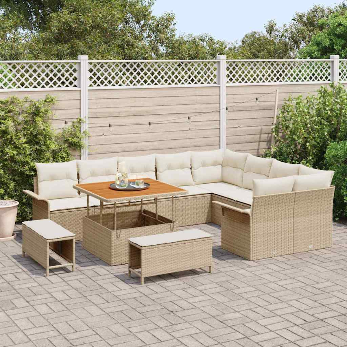 Set di mobili da esterno in vimini con cuscini beige, un tavolo in legno e due panche su un patio in pietra, contro una recinzione in legno.