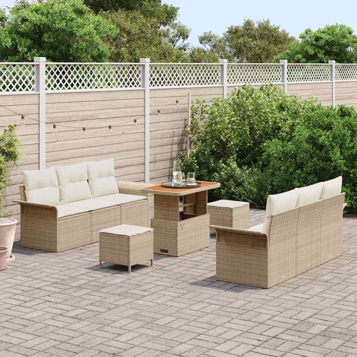 Set da patio all'aperto con divani in vimini beige, tavolo e pouf. Una recinzione bianca e una vegetazione lussureggiante formano lo sfondo.