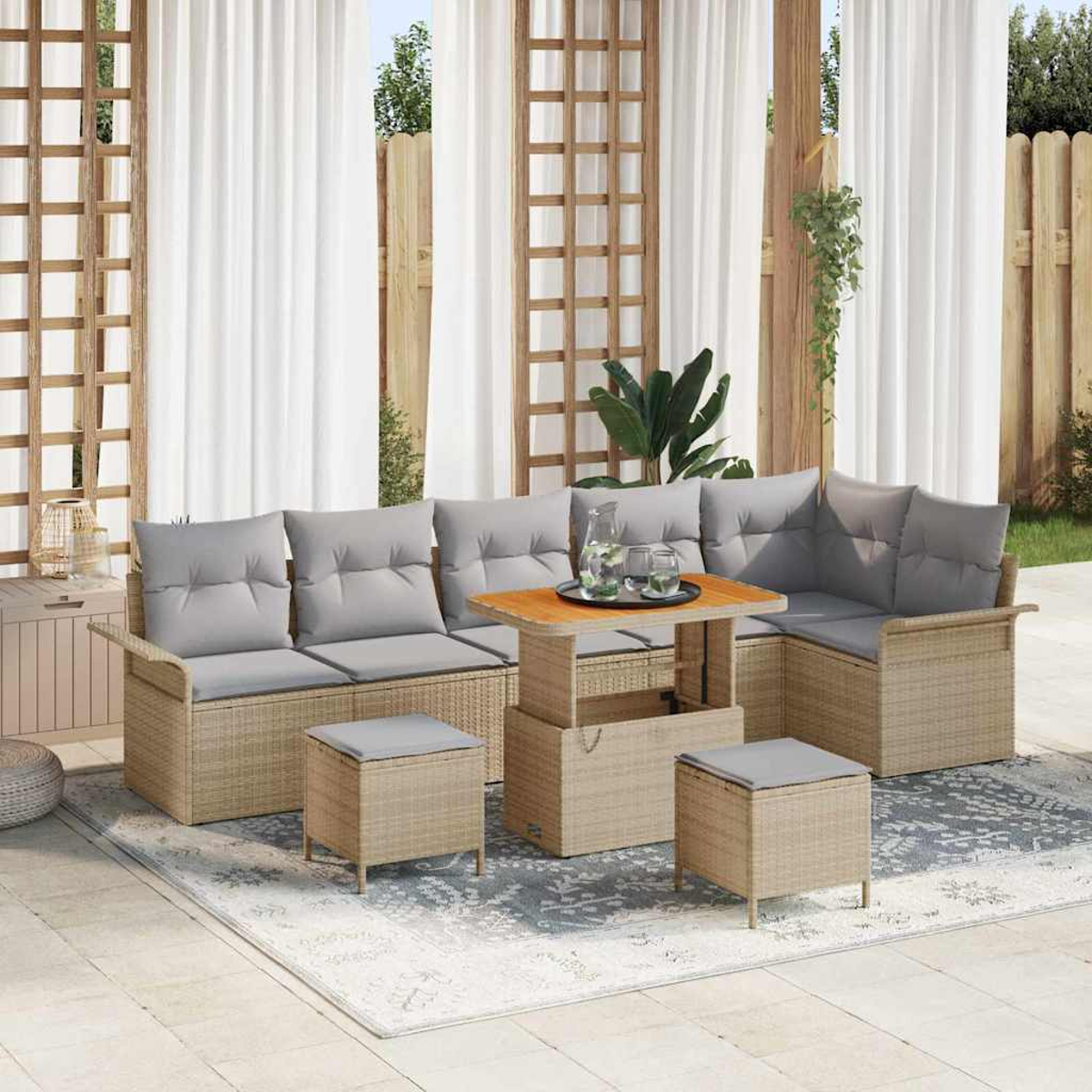 Garten-Rattan-Sofagarnitur mit grauen Kissen, Holztisch und zwei Hockern auf einem gemusterten Teppich, in der Nähe weißer Vorhänge.