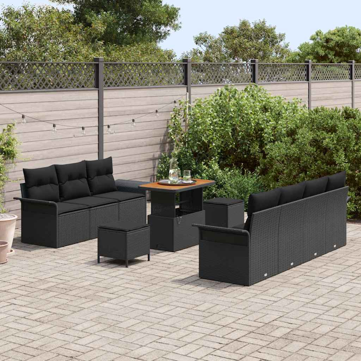 Garten-Set mit zwei schwarzen Sofas, einem Tisch und zwei Ottomanen auf einer Steinterrasse. Im Hintergrund ein Holzzaun und Büsche.