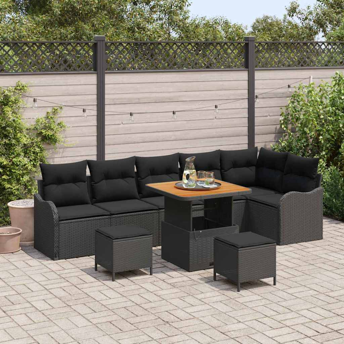 Garten-Sitzgruppe mit schwarzen Kissen, einem Tisch mit Getränken und zwei kleinen Ottomanen auf einer Terrasse. Im Hintergrund ein Zaun und Grün.