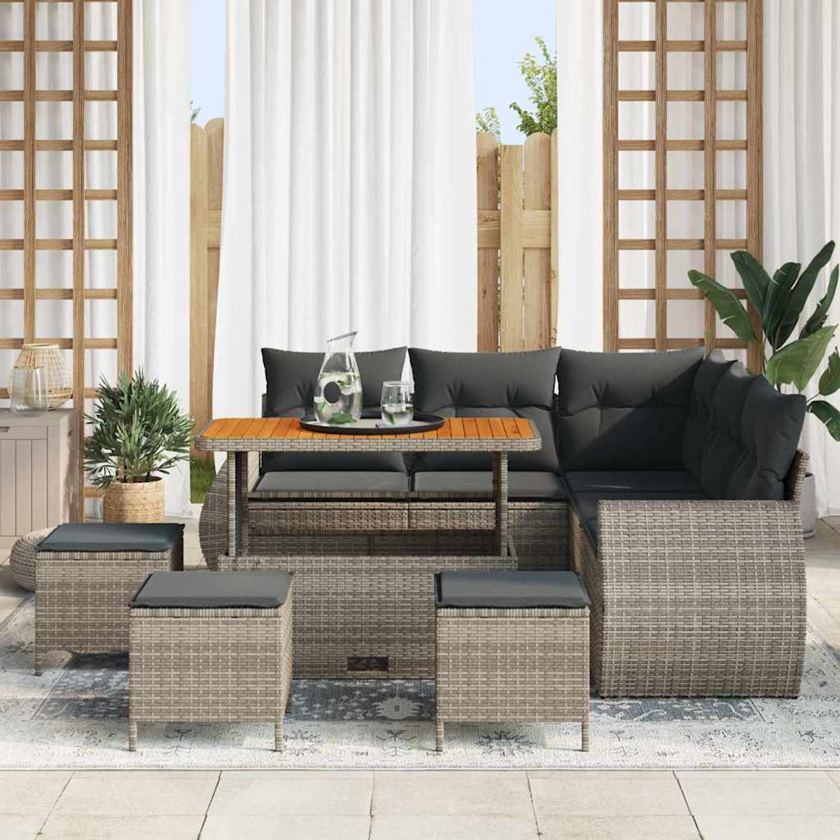 Gartensitzgruppe mit grauen Kissen, einem Holztisch und Hockern. Auf dem Tisch stehen ein Krug und ein Glas. Die Szene spielt auf einer Terrasse.