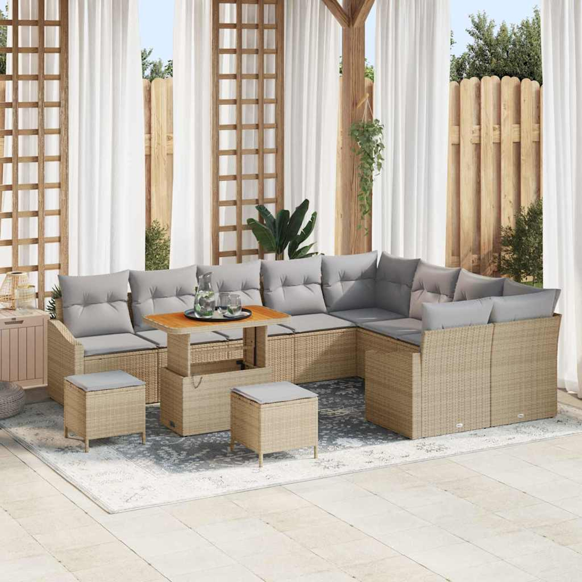 Outdoor-Wohnset mit beigen Korbmöbeln, grauen Kissen, Holztisch und zwei Hockern auf einem Teppich, Vorhängen und Holzzaun.