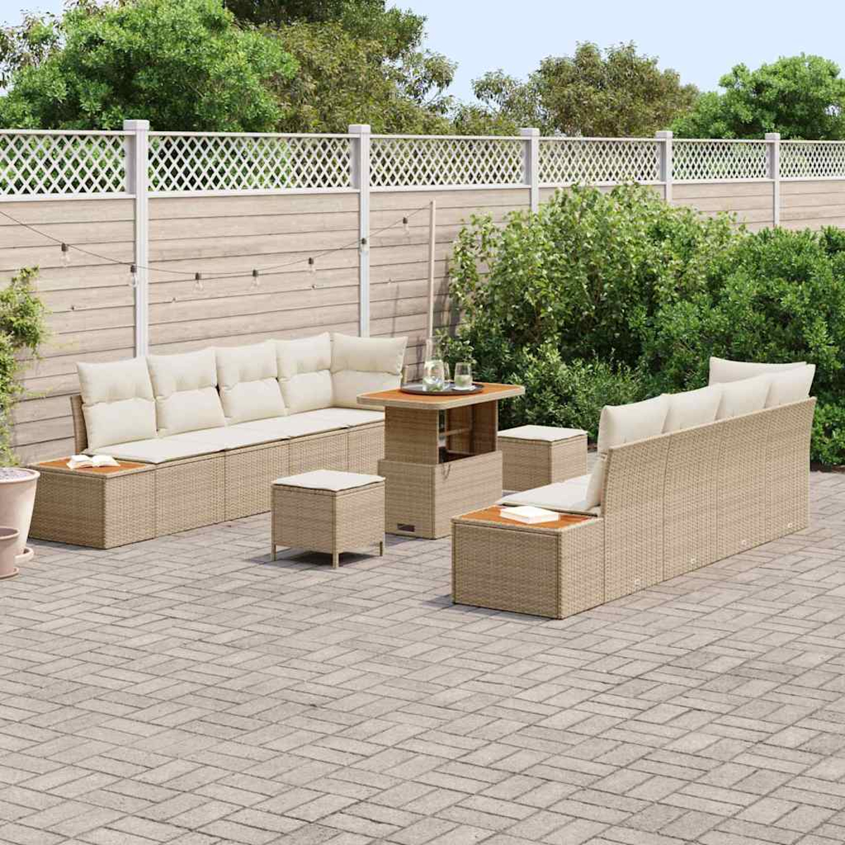 Terrassenmöbel-Set. Korbmöbel mit cremefarbenen Kissen. Tisch und Hocker auf einer Ziegelterrasse. Weißer Zaun und grüne Sträucher im Hintergrund.