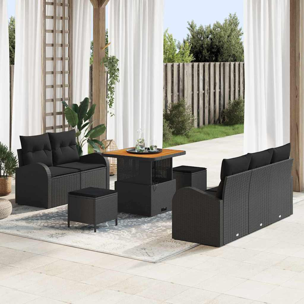 Outdoor-Terrassenset mit schwarzem Korbmöbeln, einem Tisch und einem Teppich. Weiße Vorhänge rahmen die Aussicht auf einen Holzzaun und grüne Bäume ein.