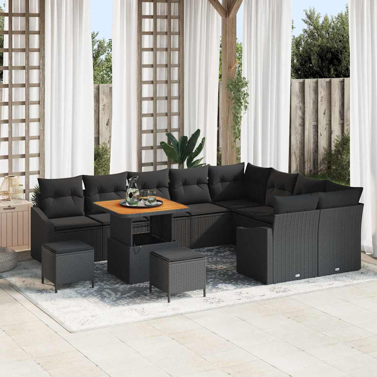 Terrassenset mit schwarzem Korbmöbel, Holztisch, Kissen und Ottomanen. Weiße Vorhänge und Holzgitter im Hintergrund.