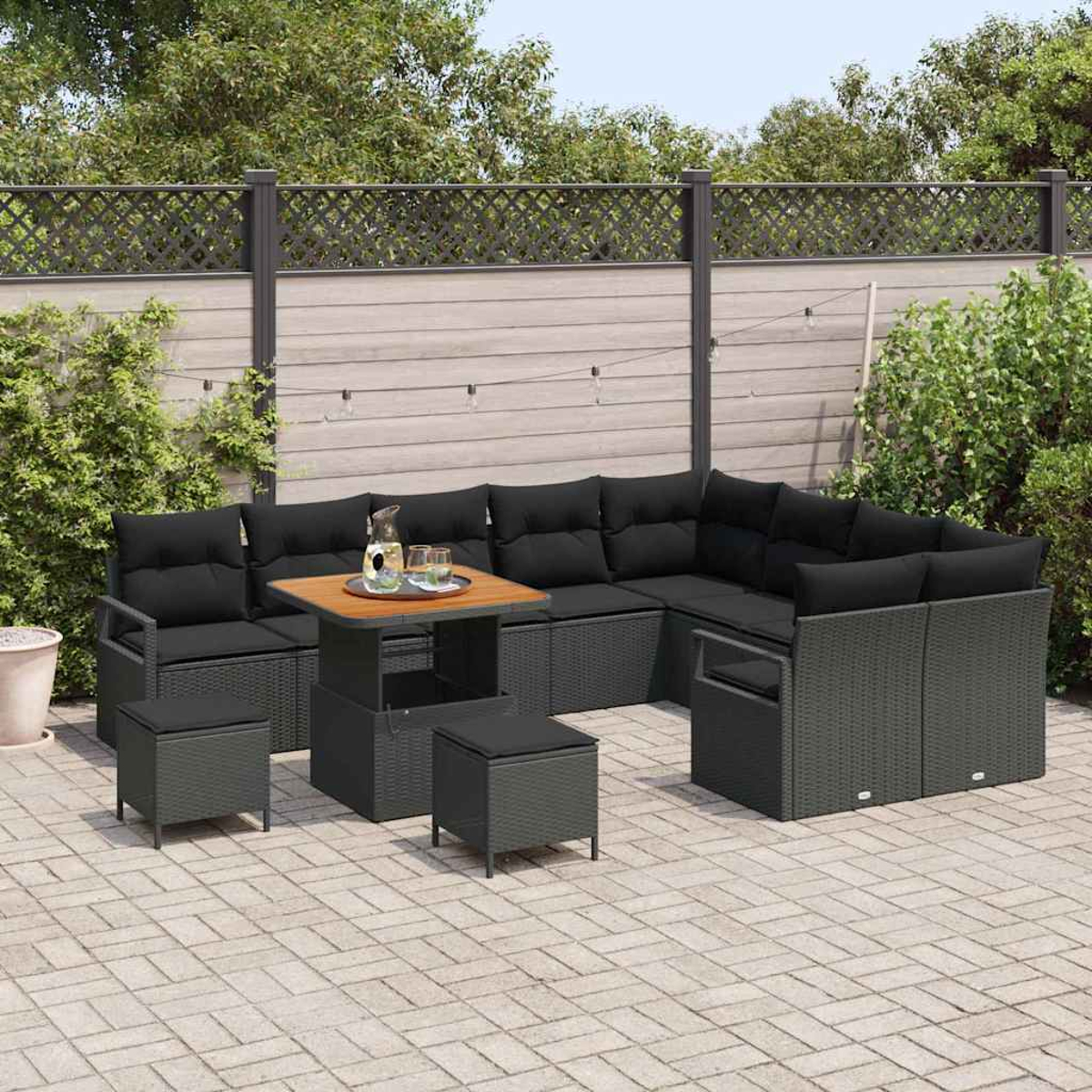 Terrassenset mit schwarzen Kissen und einem Holztisch. Graue Korbmöbel auf einer gepflasterten Terrasse, im Hintergrund ein Holzzaun.