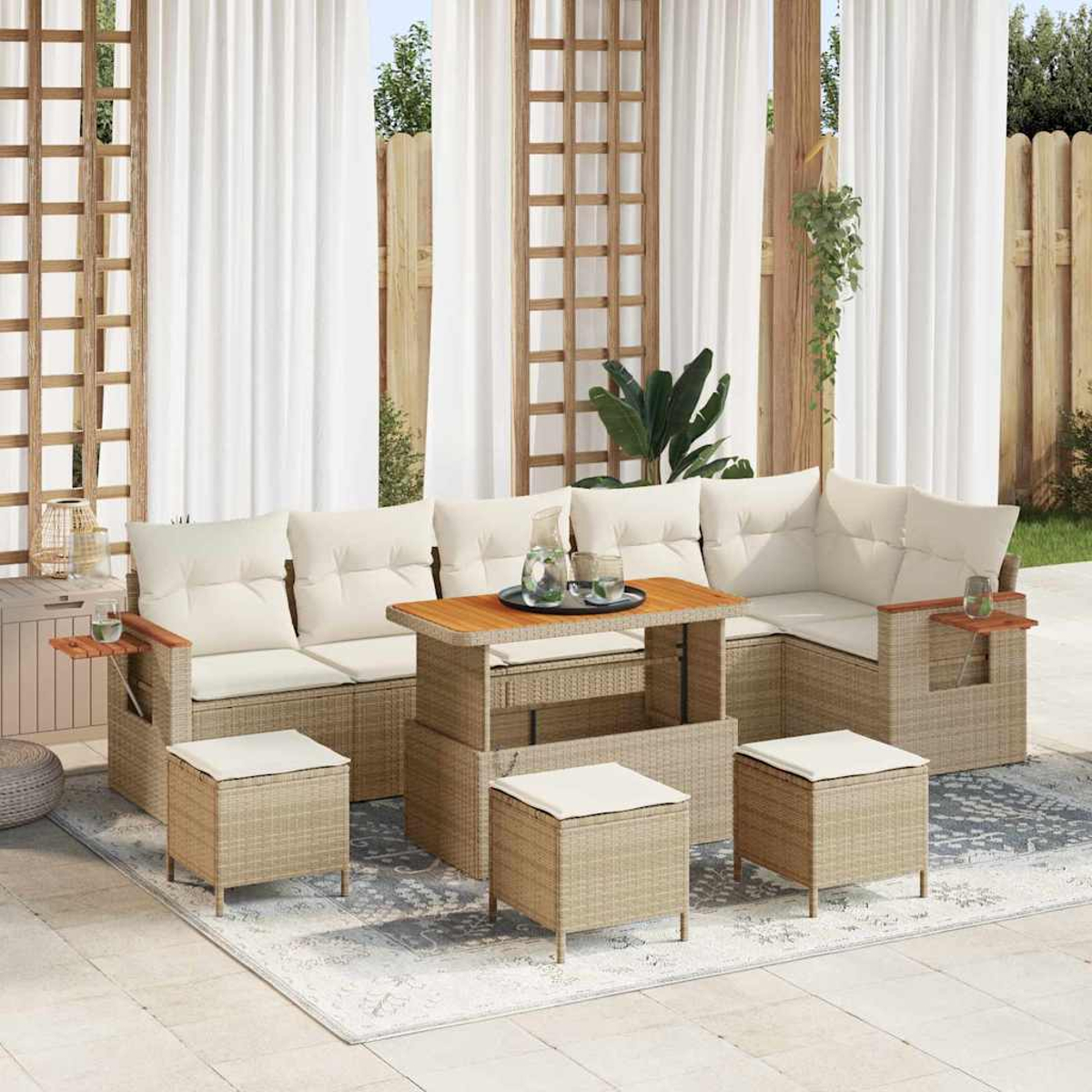 Set di mobili da esterno in vimini: un divano componibile, pouf e un tavolo, su un tappeto, con tende bianche e una staccionata in legno sullo sfondo.