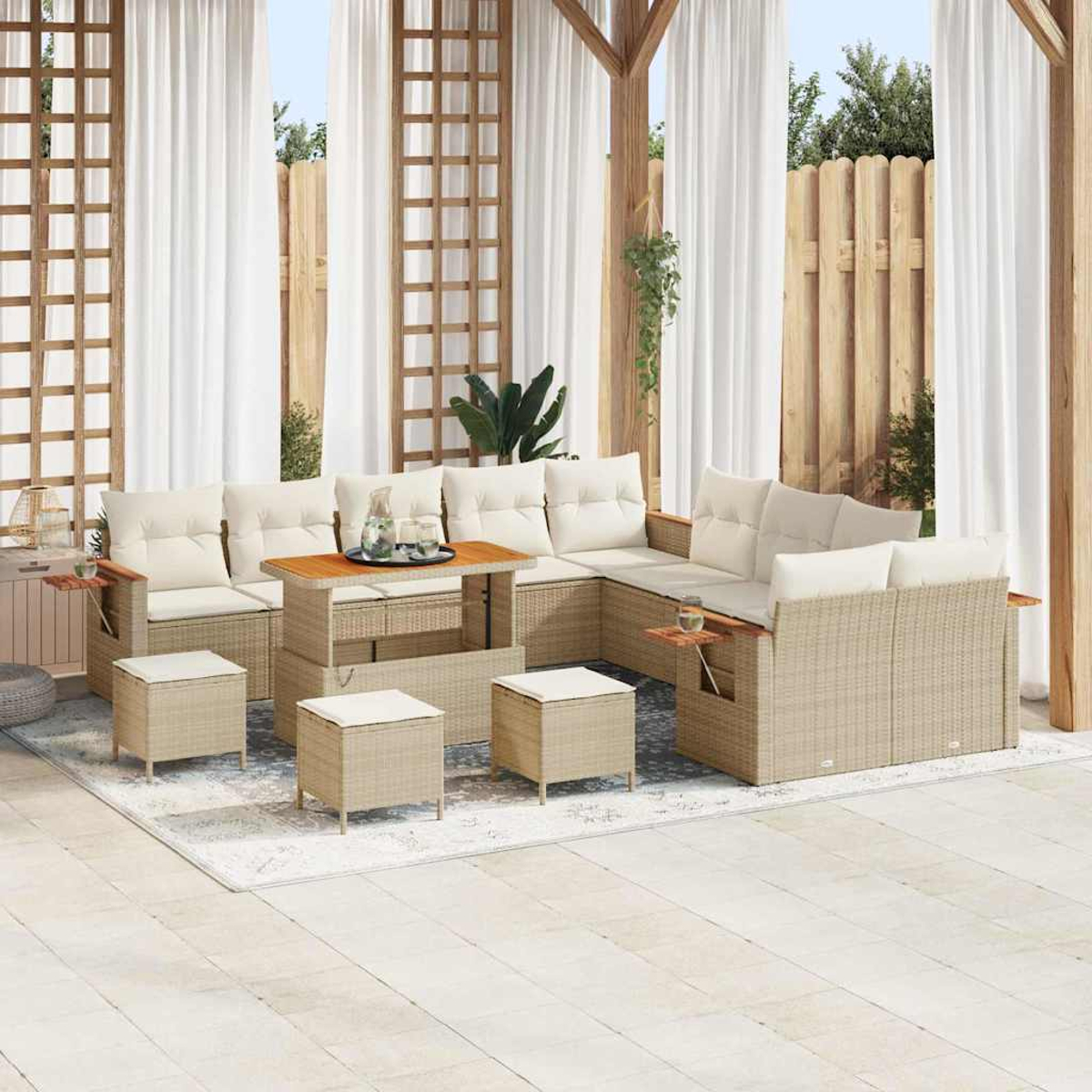 Divano angolare in vimini beige da esterno con cuscini bianchi, tavolino in legno e pouf abbinati. Sfondo con tende bianche e recinzione in legno.