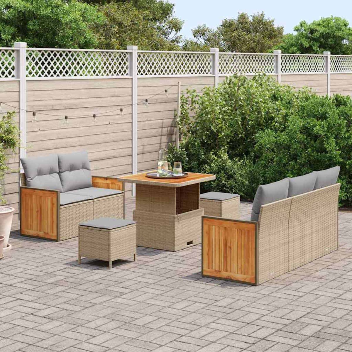 Set da patio esterno con mobili in vimini beige, cuscini grigi e un tavolo in legno. Circondato da una recinzione e vegetazione.