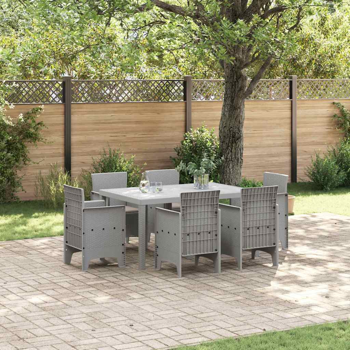 Scena all'aperto con tavolo e sedie grigie. Una staccionata in legno e vegetazione circondano il patio, pavimentato con mattoni.