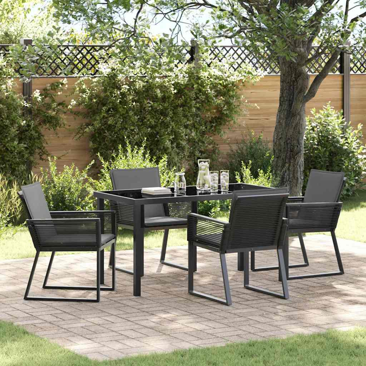 Essgruppe im Freien auf einer Ziegelterrasse. Ein quadratischer Tisch mit vier Stühlen, Gläsern und einer Kanne in einem Garten.