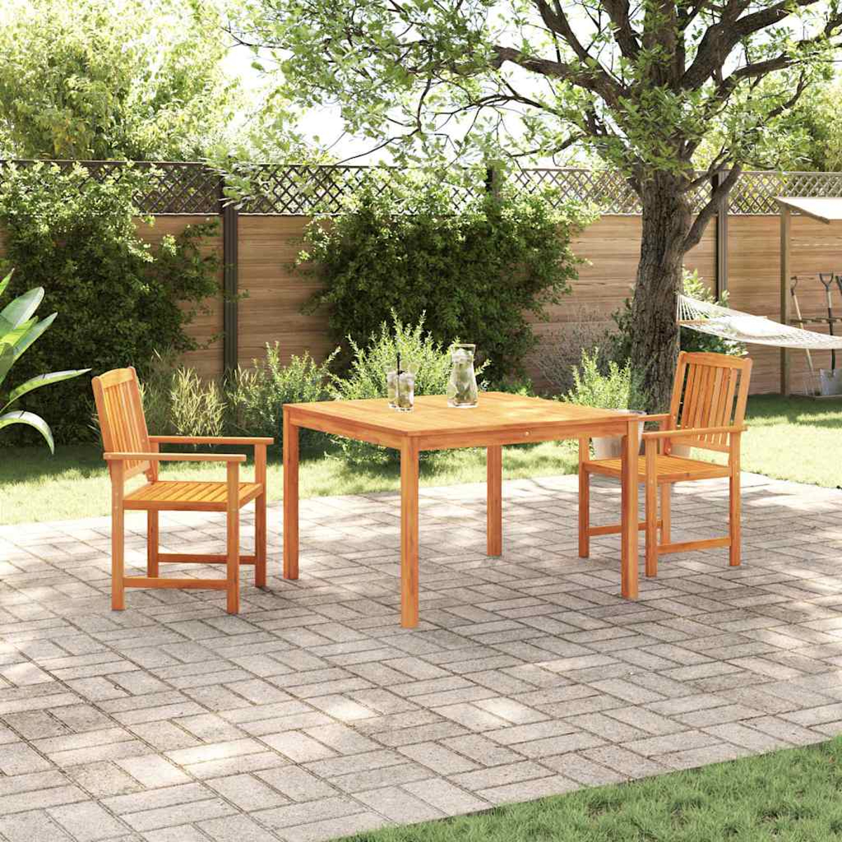 Ein Holztisch mit zwei Stühlen auf einer Terrasse. Im Hintergrund hängt eine Hängematte, ringsherum Bäume und Grün.