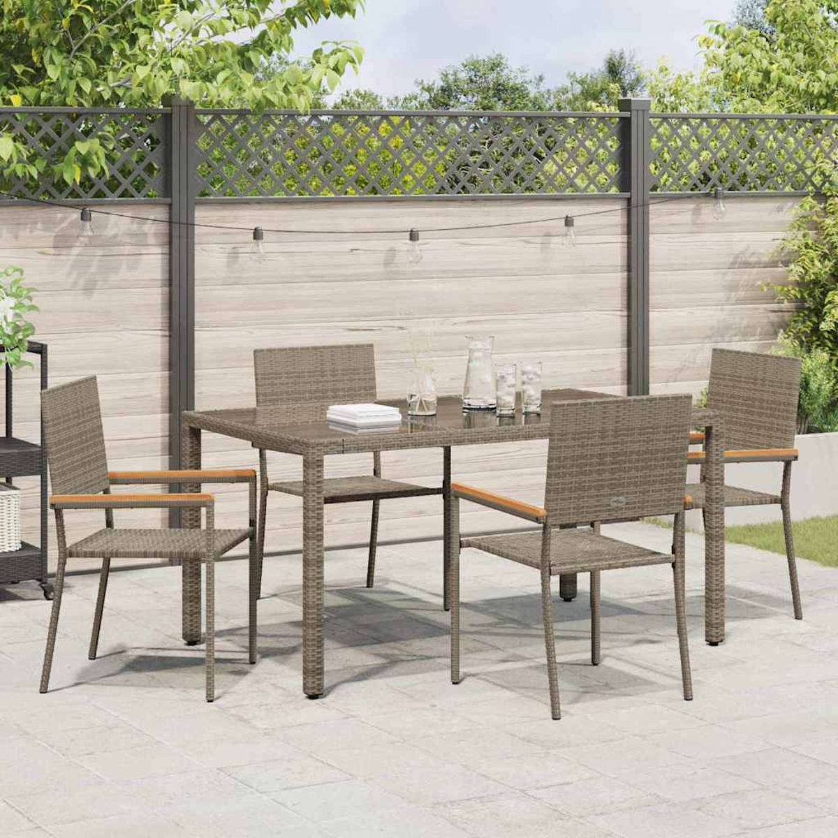 Essgruppe im Freien: Tisch, vier Stühle auf einer Terrasse. Im Hintergrund ein Zaun und Grün. Auf dem Tisch stehen Gläser und eine Kanne.