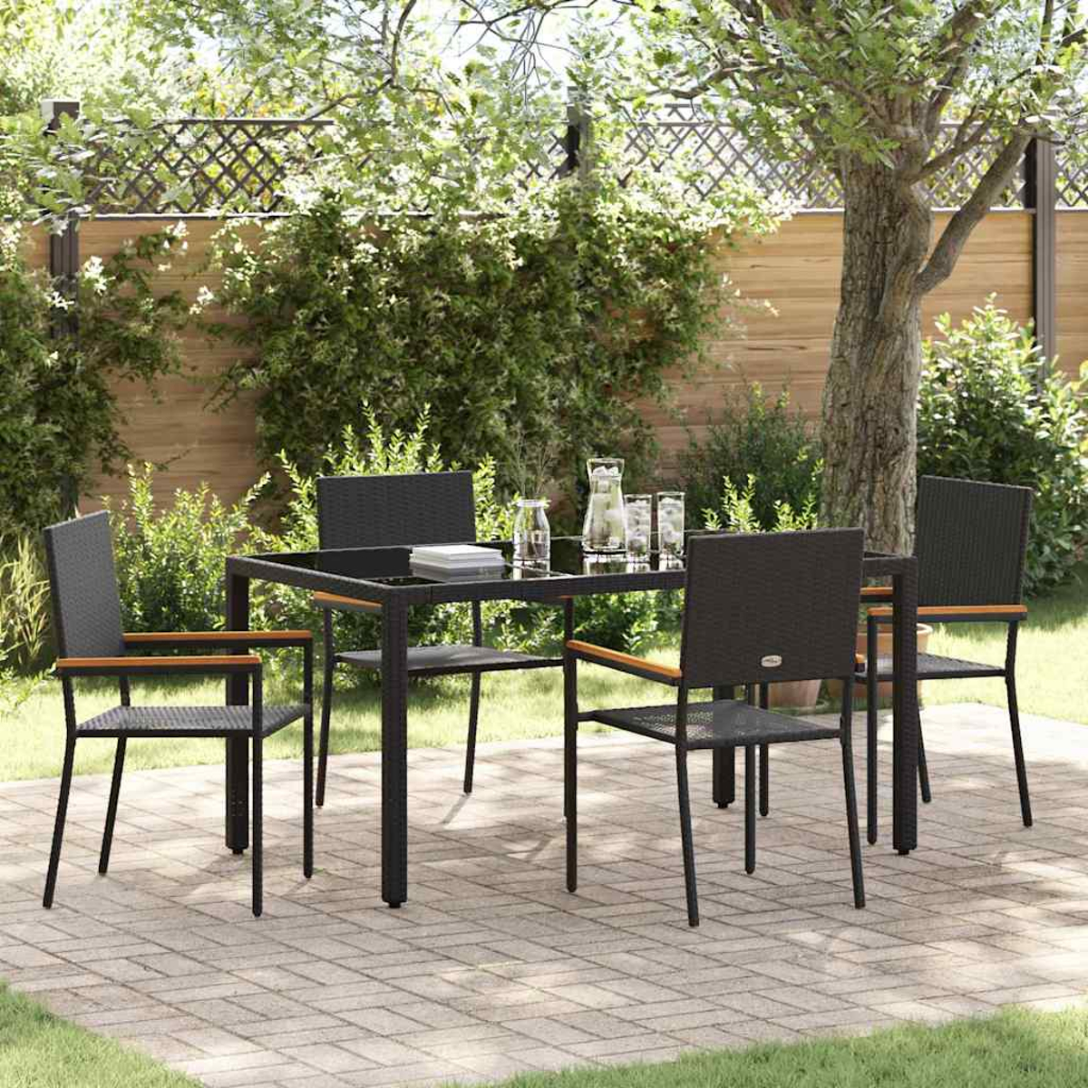 Essgruppe im Freien mit schwarzem Tisch und Stühlen auf einer gepflasterten Terrasse. Grüne Pflanzen, Bäume und ein Holzzaun im Hintergrund.