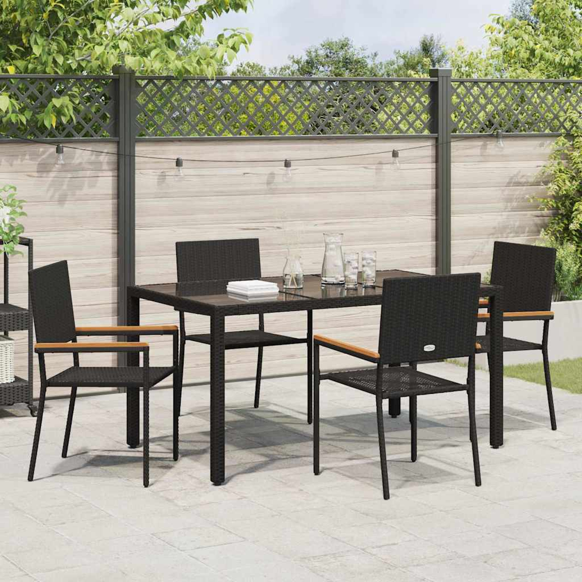 Ein schwarzes Ess-Set für den Außenbereich mit Tisch und vier Stühlen. Im Hintergrund ein Zaun und Grün. Gläser und eine Kanne auf dem Tisch.