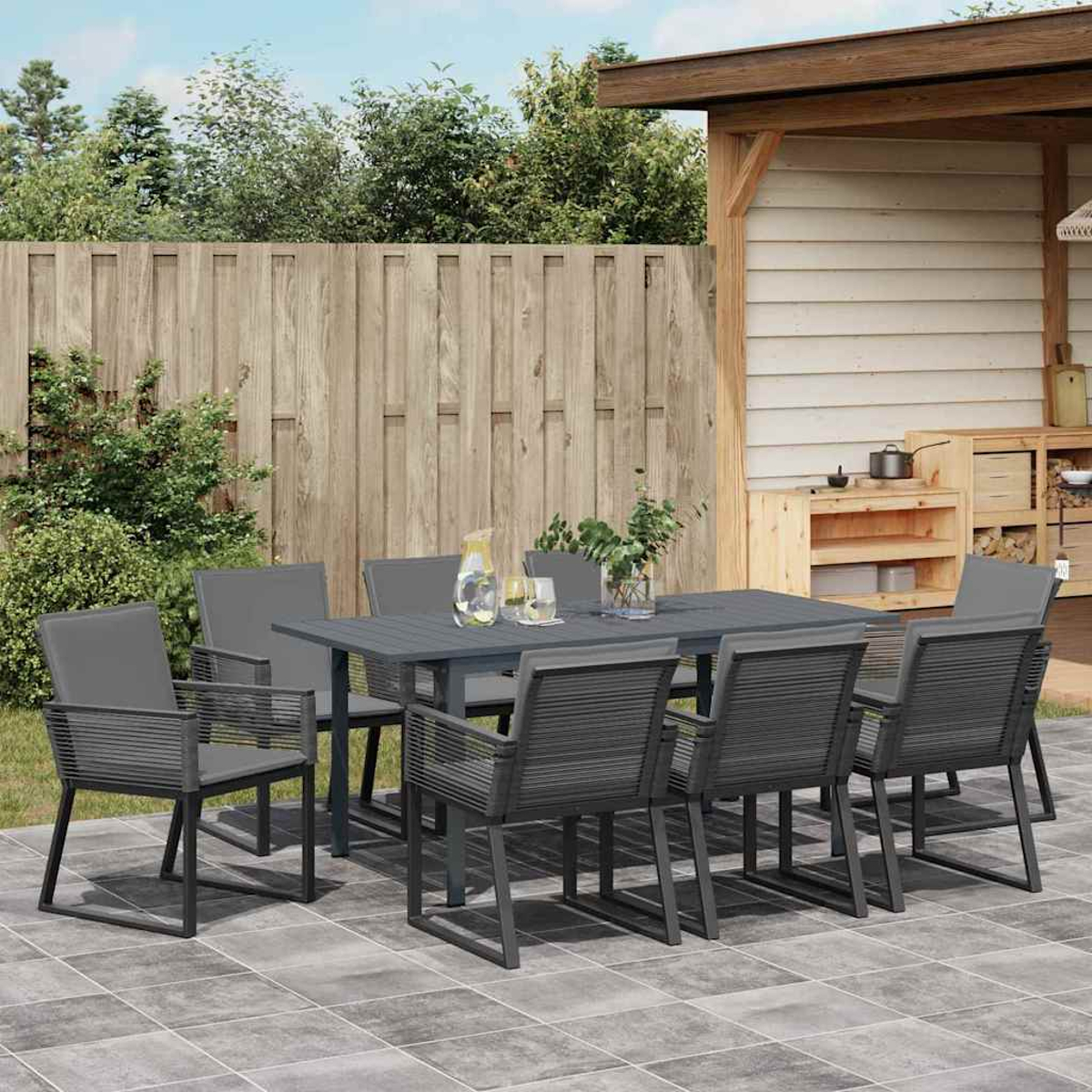 Essgruppe im Freien: grauer Tisch und Stühle auf der Terrasse. Holzzaun und Schuppen im Hintergrund. Pflanzen und Gläser auf dem Tisch.