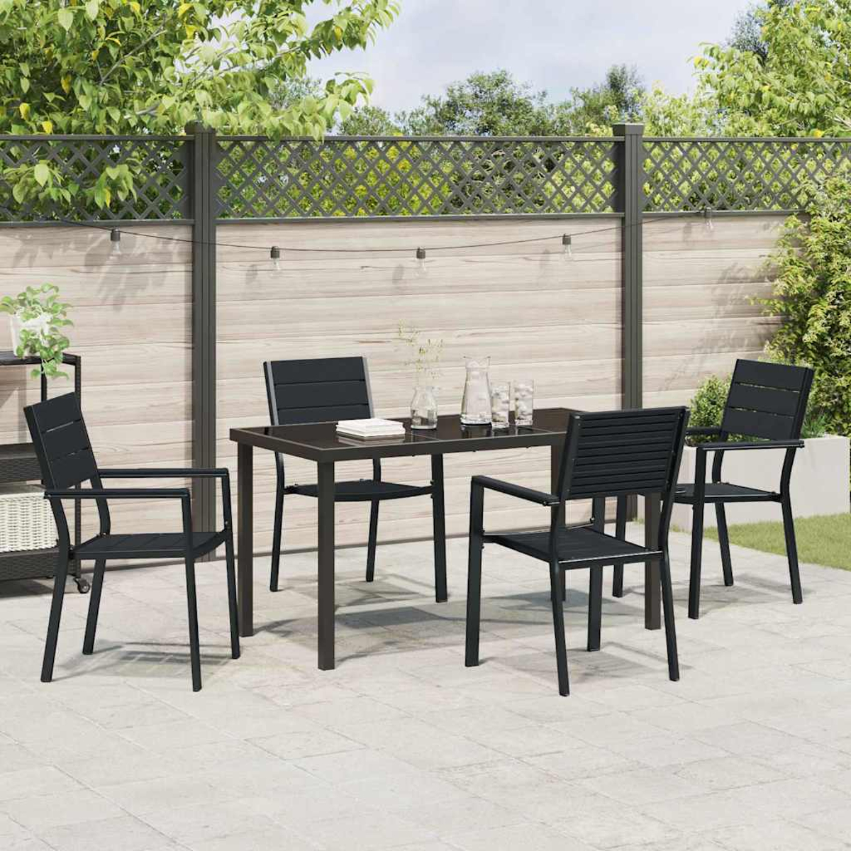 Essgruppe im Freien mit einem schwarzen Tisch und vier schwarzen Stühlen auf einer Terrasse. Im Hintergrund ein Holzzaun und Bäume.