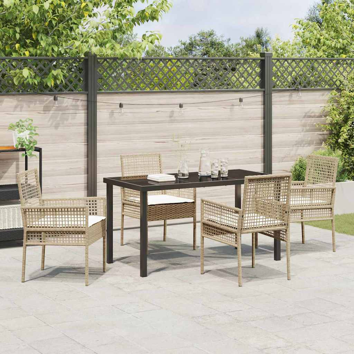 Set da pranzo all'aperto. Tavolo nero, sedie in vimini con cuscini bianchi. Ambiente giardino con recinzione e verde.