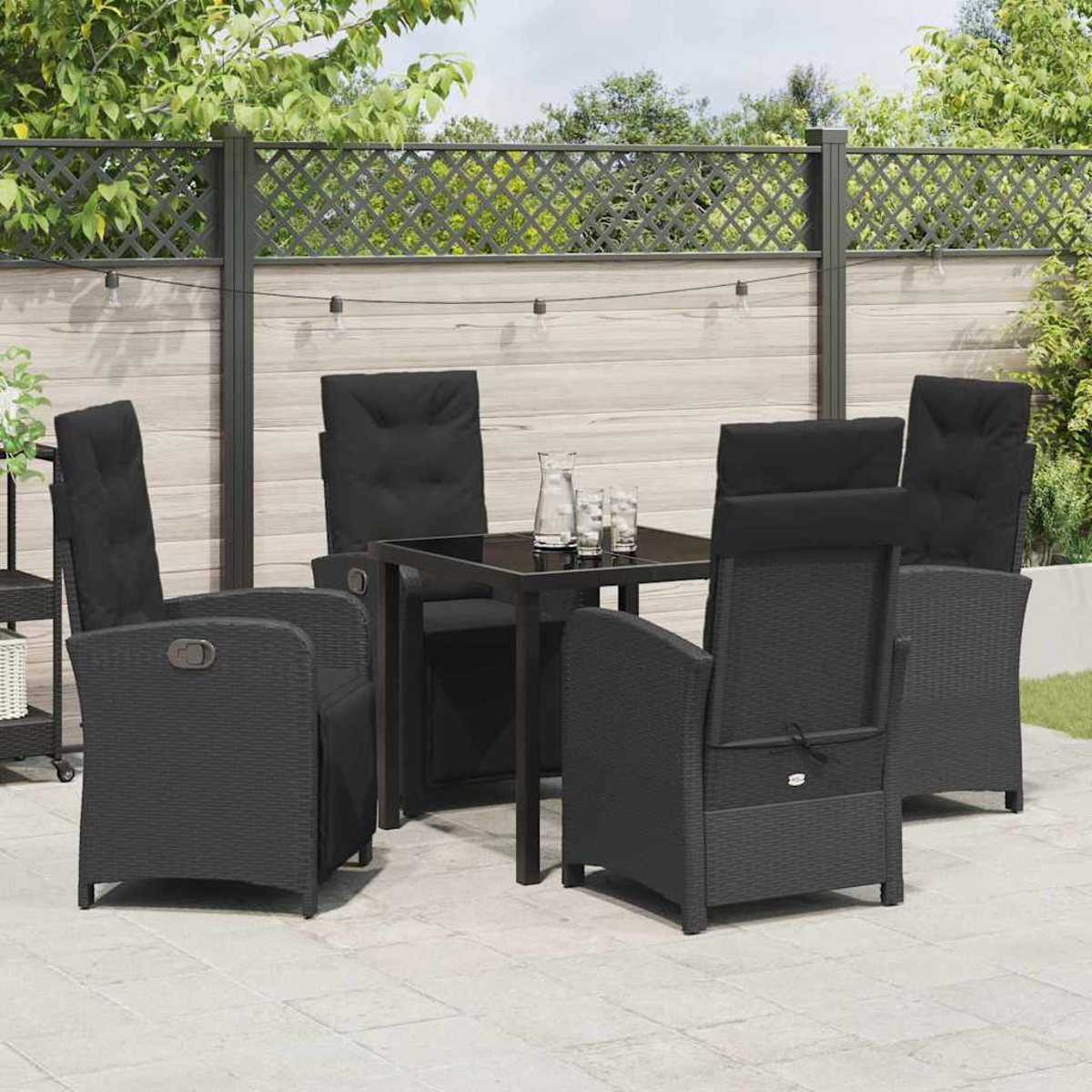 Essgruppe im Freien: schwarze Stühle und Tisch auf einer Terrasse. Hintergrund: Zaun, Bäume und Lichterketten.