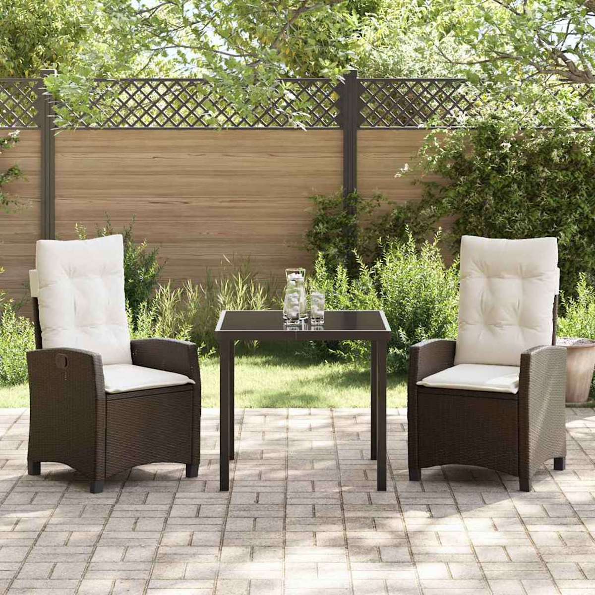 Zwei braune Korbstühle mit weißen Kissen und ein kleiner Tisch auf einer Terrasse. Im Hintergrund ein Holzzaun und Grün.