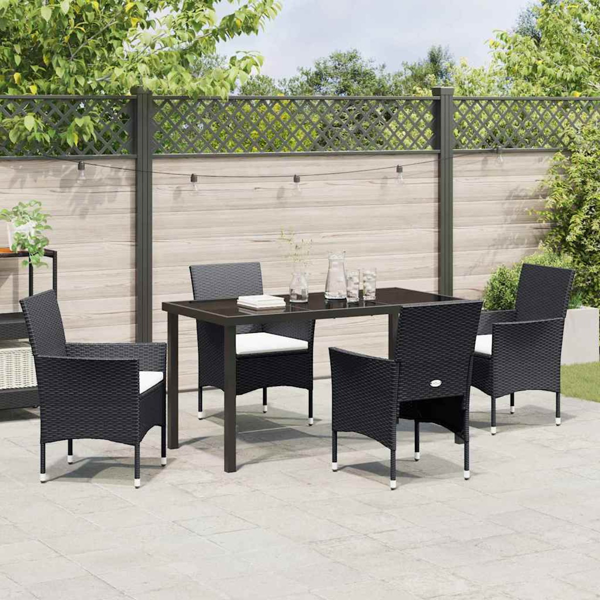 Set da patio esterno con tavolo e sedie scure. Una recinzione e vegetazione sullo sfondo. C'è luce solare.