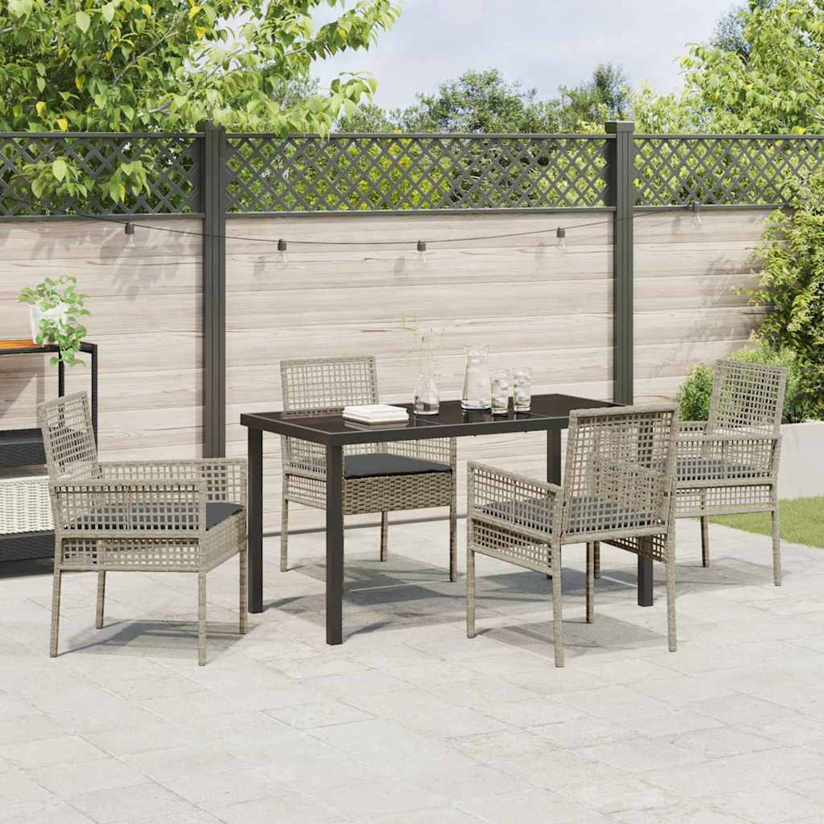 Set da pranzo all'aperto. Un tavolo nero con quattro sedie, posto su un patio in pietra. Recinzione in legno e vegetazione sullo sfondo.