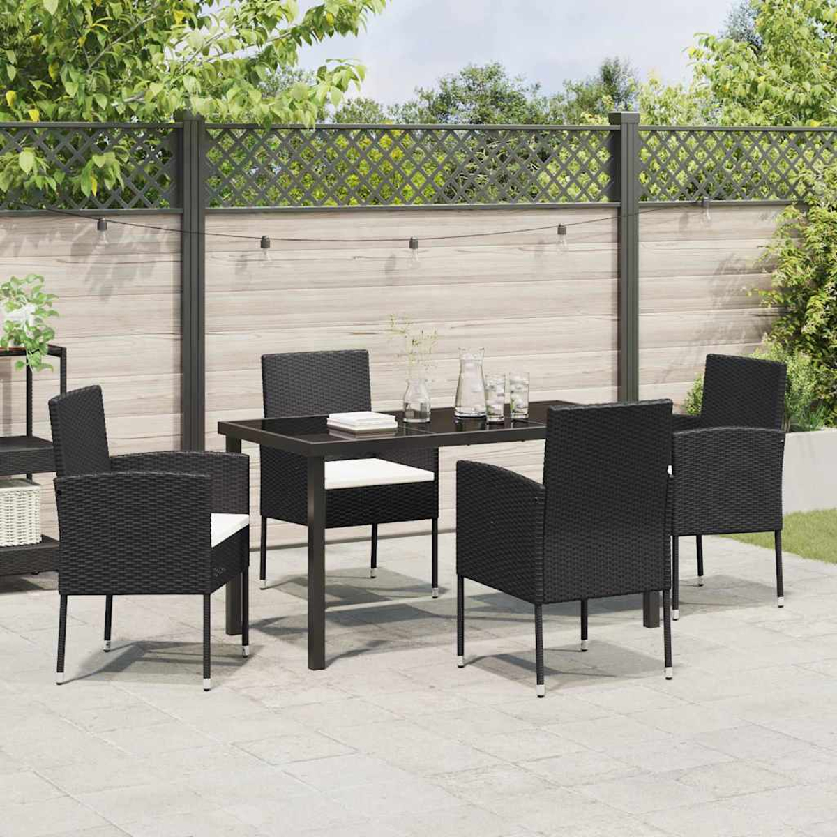 Szene im Freien: Schwarze Korbstühle um einen schwarzen Tisch mit Gläsern. Im Hintergrund ein Zaun und Laub.