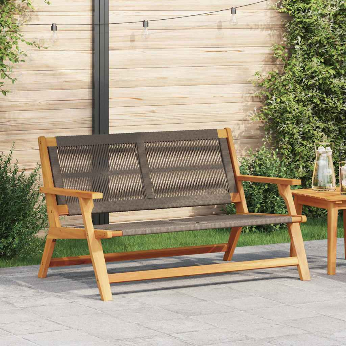 Banco de madera con respaldo y asiento tejidos sobre un patio. Follaje verde, valla de madera y luces de cadena en el fondo.