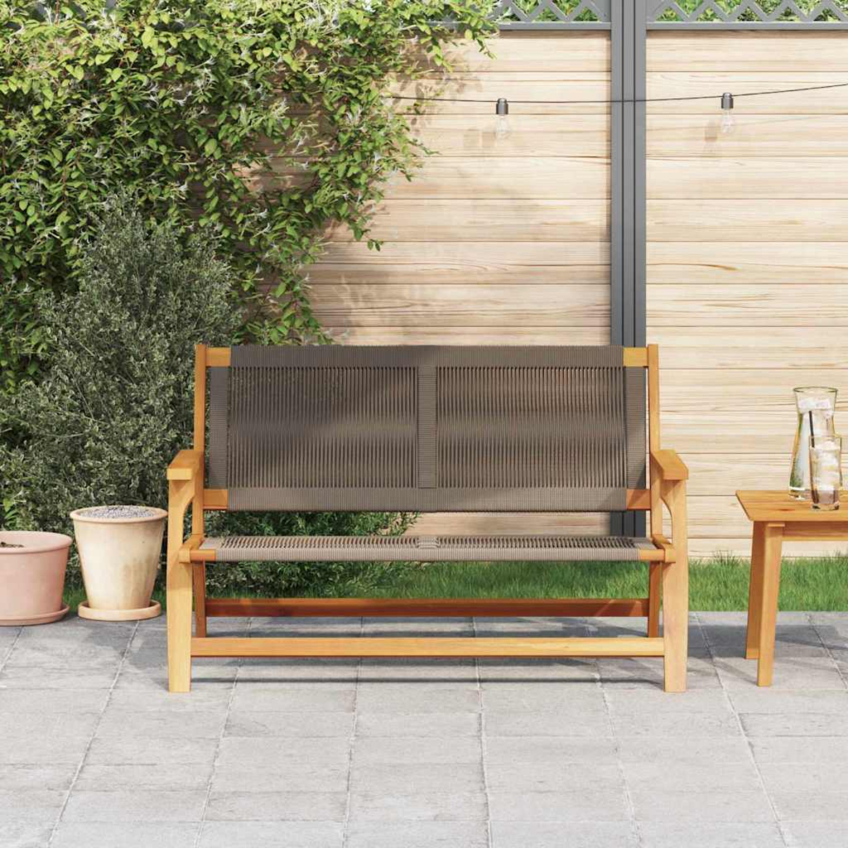 Banqueta de madera con respaldo y asiento tejido gris en el patio. Follaje verde, valla de madera y mesa pequeña con jarra de agua al fondo.