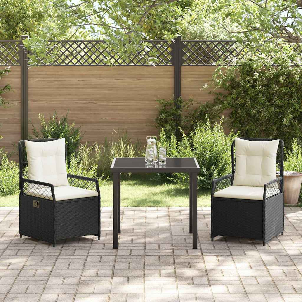 Terrassenset mit zwei schwarzen Korbstühlen, cremefarbenen Kissen, einem kleinen Tisch und Gläsern. Holzzaun und Grün im Hintergrund.