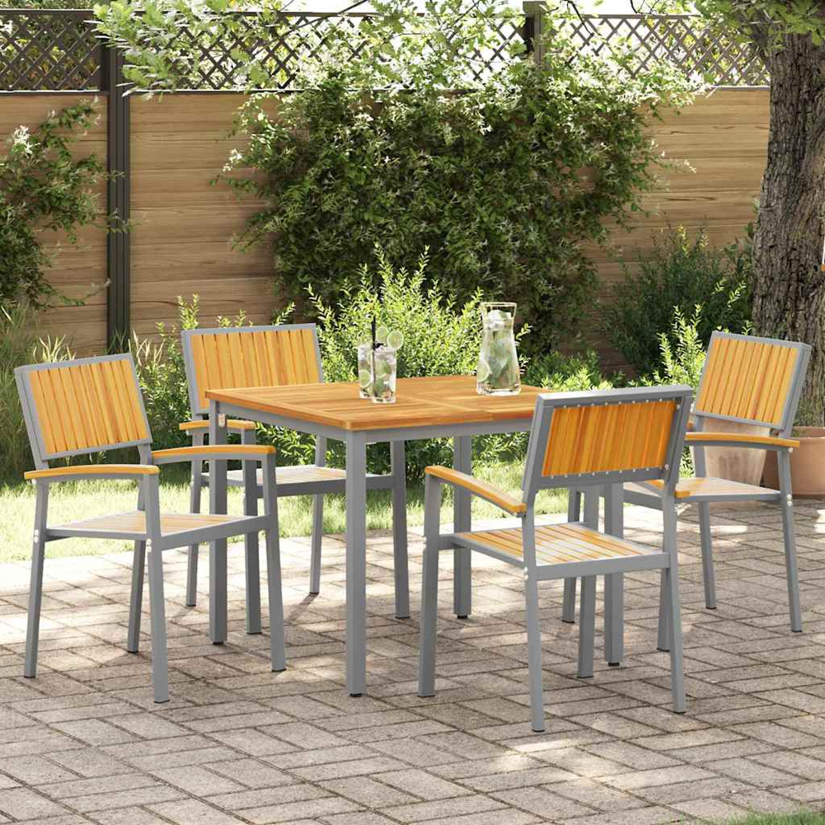 Gartenarrangement mit einem quadratischen Holztisch und vier Stühlen. Getränke stehen auf dem Tisch. Grünes Laub und ein Holzzaun im Hintergrund.