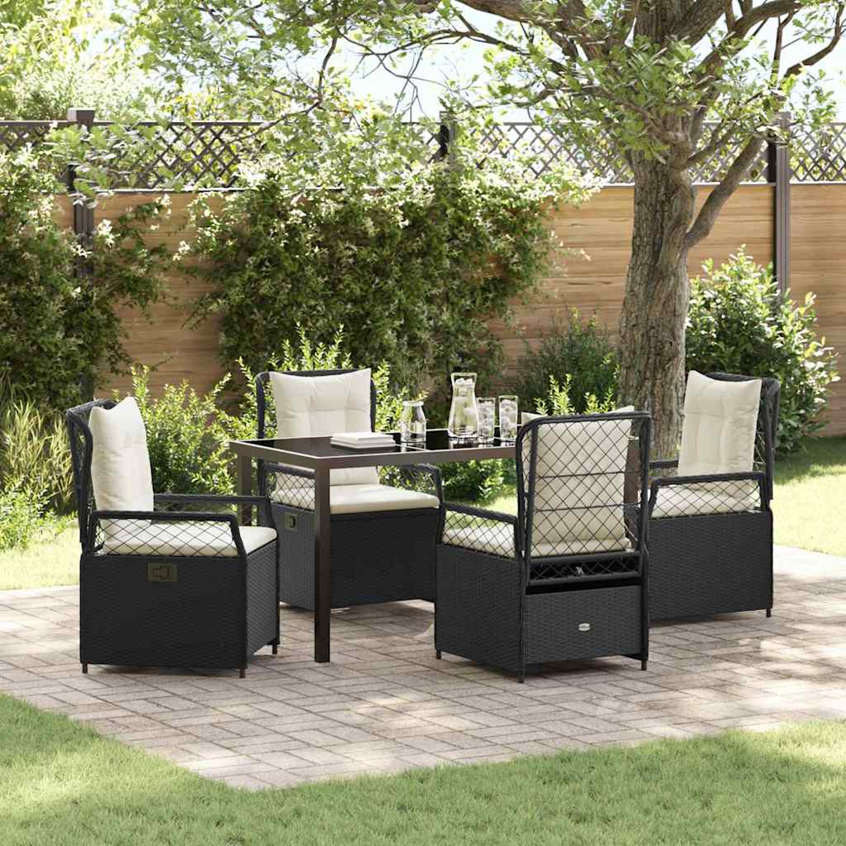 Terrassenset mit schwarzen Korbstühlen, cremefarbenen Kissen und Glastisch. Grünes Laub und Holzzaun im Hintergrund.