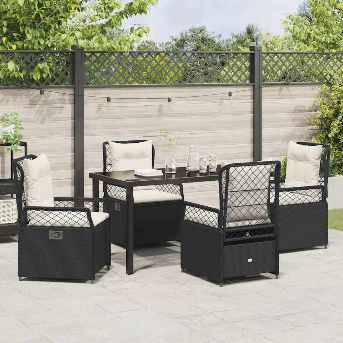 Terrassenset mit schwarzen Korbstühlen, cremefarbenen Kissen, Tisch und dekorativem Zaun im Hintergrund. Pflanzen und Lichter sind vorhanden.