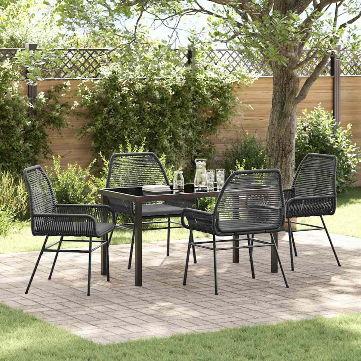 Set da pranzo all'aperto: sedie e tavolo neri su un patio in mattoni, con un albero e vegetazione sullo sfondo.
