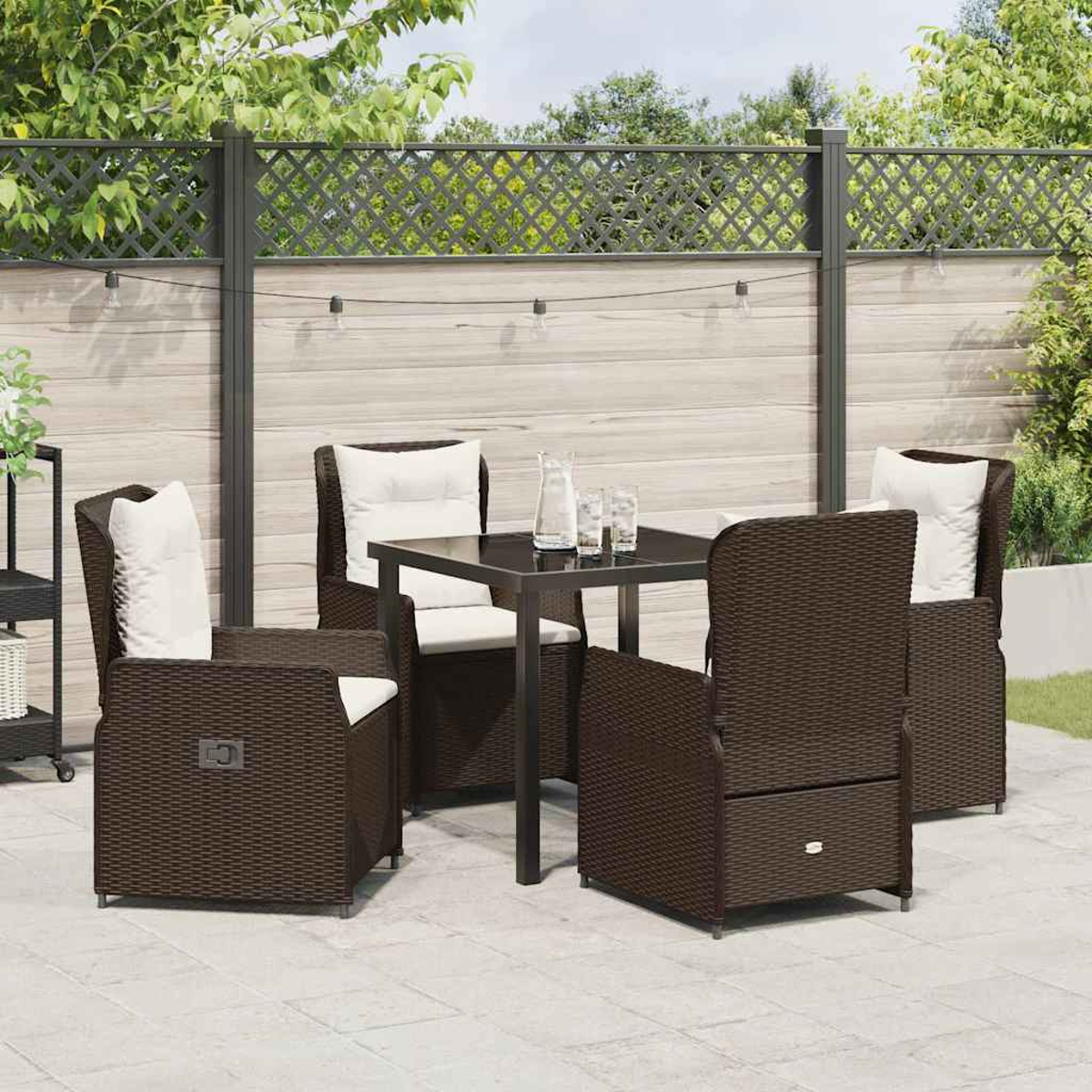 Terrassengarnitur im Freien mit braunen Korbstühlen, weißen Kissen, einem dunklen Tisch und einem Zaun im Hintergrund. Gläser und eine Kanne stehen auf dem Tisch.