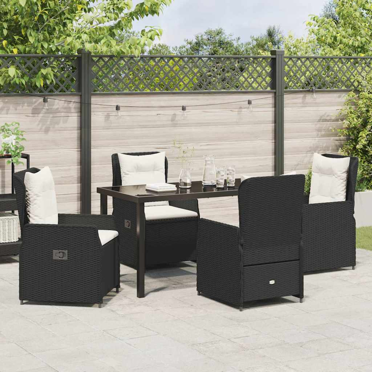 Garten-Terrassenset mit schwarzen Korbstühlen, weißen Kissen und einem schwarzen Tisch. Ein Holzzaun und Grün im Hintergrund.