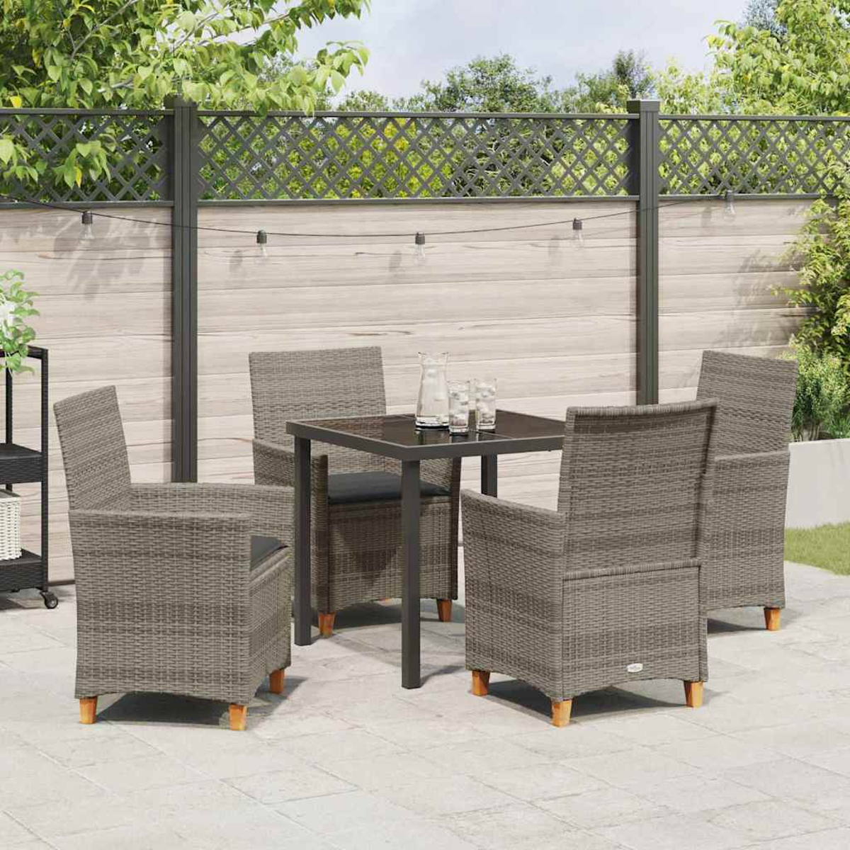 Terrassenset im Freien mit vier grauen Korbstühlen um einen quadratischen Tisch. Ein Holzzaun und Bäume sind im Hintergrund.
