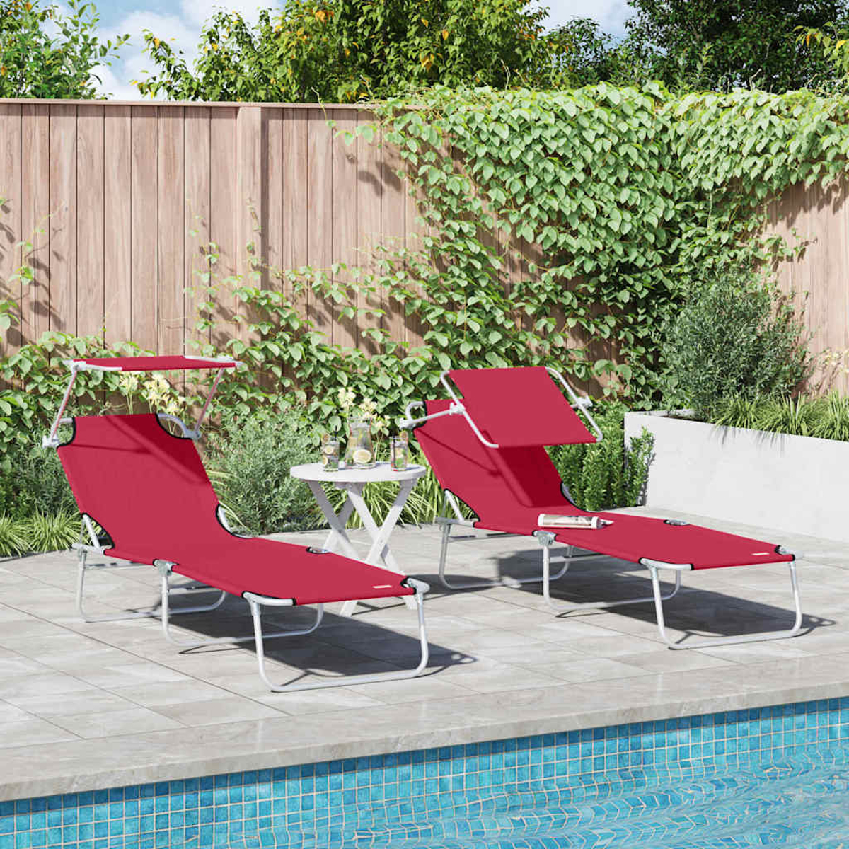 Zwei rote Liegestühle mit Sonnenschirmen am Pool. Ein kleiner Tisch mit Getränken steht dazwischen. Hintergrund: Holzzaun, Grün.