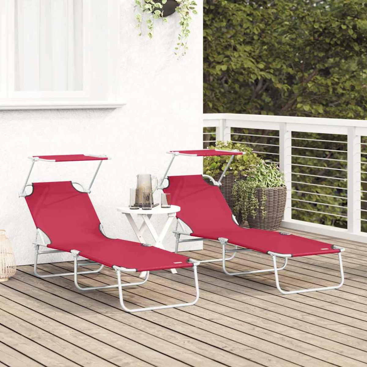 Zwei rote Liegestühle mit weißen Gestellen stehen auf einer Holzterrasse. Ein kleiner Tisch mit Krug und Gläsern steht dazwischen. Grünes Laub im Hintergrund.