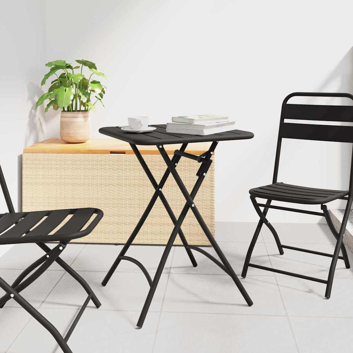 Un juego de mesa y sillas plegables negras sobre un suelo embaldosado. Una planta, una taza y libros están sobre la mesa. Un cofre de mimbre está en el fondo.