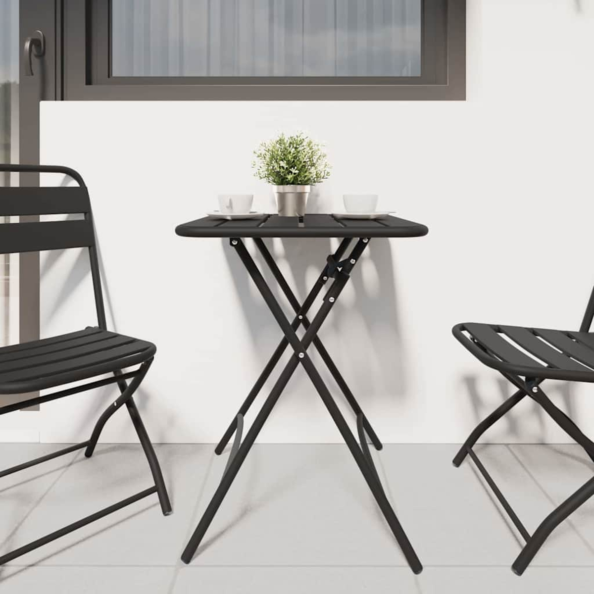 Escena de balcón con mesa y sillas plegables negras. La mesa tiene una planta pequeña y tazas. Pared blanca y ventana al fondo.