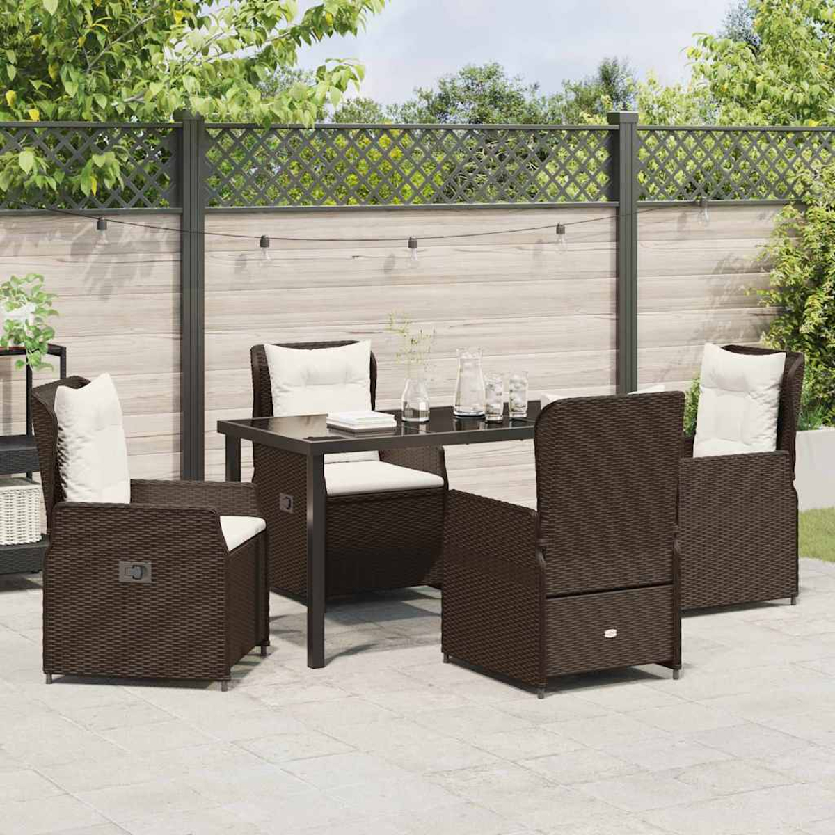 Terrassenmöbel-Set mit braunen Korbstühlen, einem Tisch und weißen Kissen. Im Hintergrund ein Zaun und Pflanzen.